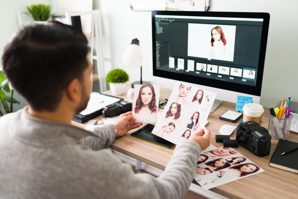 Hispanic young man seen from behind looking at some print photos and retouching some others on the computer